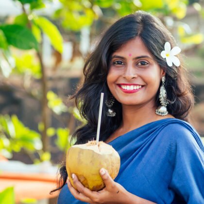 A happy Indian woman enjoying a coconut drink on a tropical beach, symbolizing a light yet enriching immersion into the Hindi language.