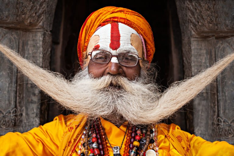 An elderly Indian man with a long white mustache and beard, wearing a vibrant orange turban and traditional spiritual markings on his forehead, symbolizing India’s sacred and spiritual heritage.